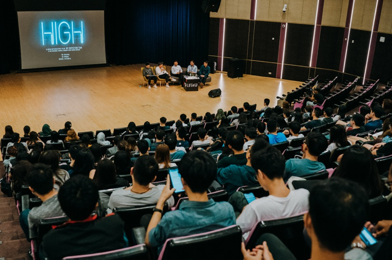 A large audience watches a presentation from speakers on a stage; "HIGH" is shown on a screen.